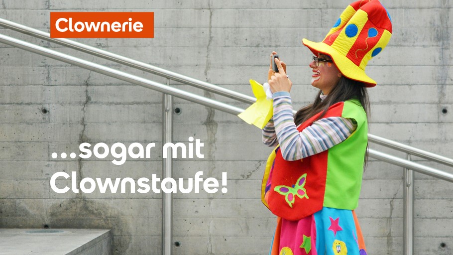 Eine Frau in Clownkostüm hält ein Telefon, steht vor einem Betongebäude mit Geländern.