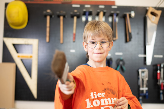 Ein Junge in einer Werkstatt hält einen Hammer und trägt eine orangefarbene T-Shirt.