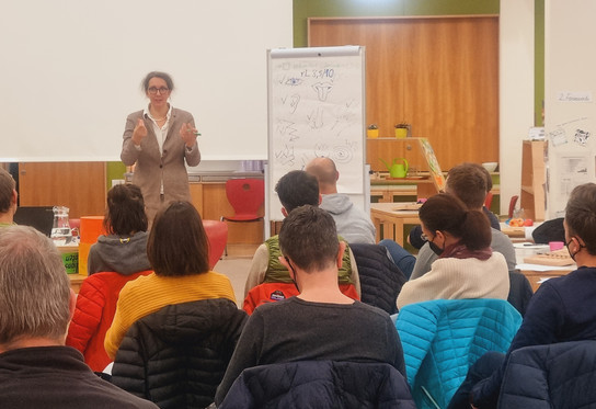 Eine Frau steht vor einem Publikum und hält eine Präsentation in einem Klassenzimmer.