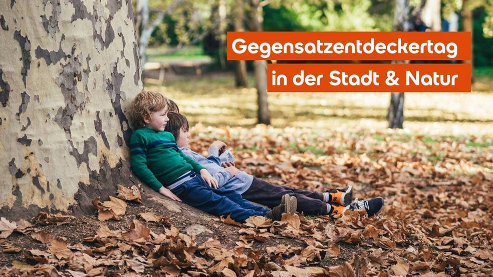Zwei Kinder sitzen an einem Baumstamm auf einem Boden mit gefallenen Blättern.